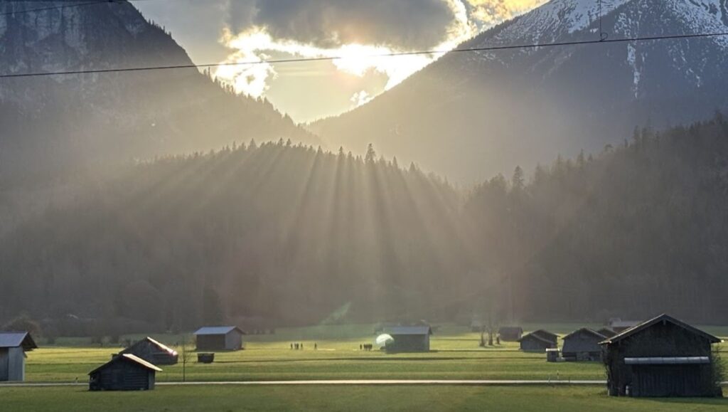 Sonnenstrahlen brechen durch Wolken über einem weiten Tal im Zugspitzland – Stadel und schneebedeckte Berggipfel im Hintergrund, goldenes Licht liegt über der Landschaft.