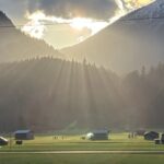 Sonnenstrahlen brechen durch Wolken über einem weiten Tal im Zugspitzland – Stadel und schneebedeckte Berggipfel im Hintergrund, goldenes Licht liegt über der Landschaft.